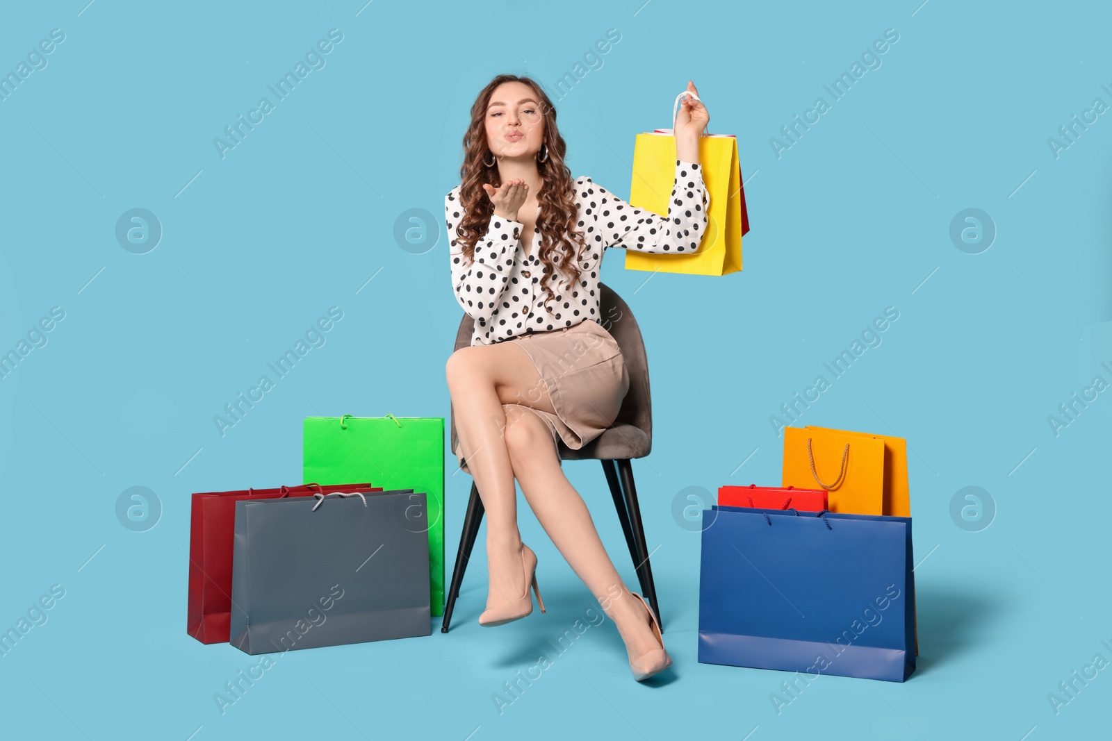 Photo of Young woman holding colorful shopping bags and blowing kiss on chair against light blue background