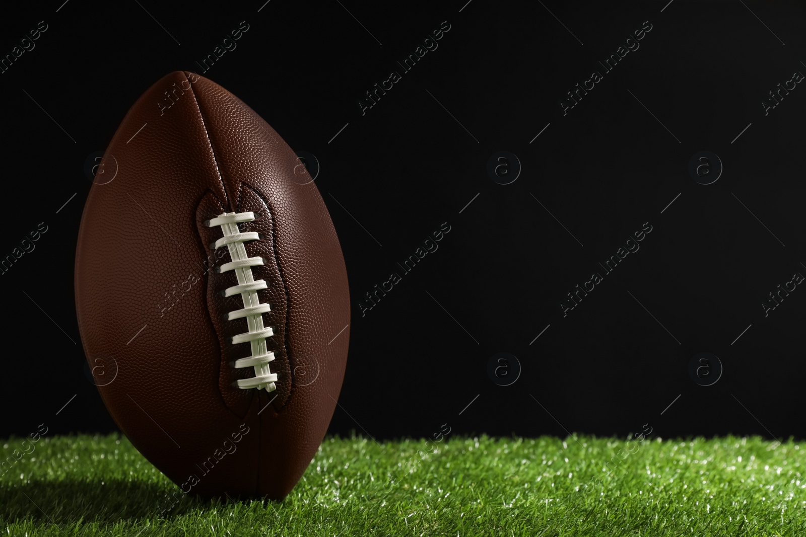 Photo of American football ball on green grass against black background. Space for text