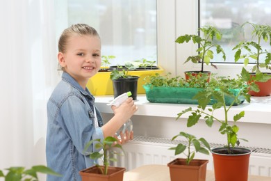 Cute little girl spraying seedlings in plastic container on windowsill indoors