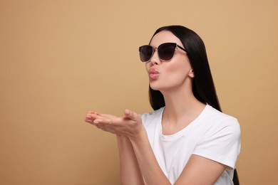 Beautiful young woman in stylish sunglasses blowing kiss on beige background. Space for text