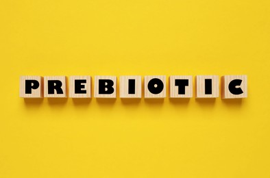 Wooden cubes with word Prebiotic on yellow background, flat lay