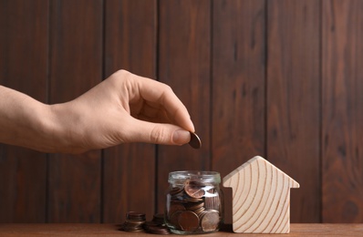 Woman putting coin into glass jar near house model on table against wooden background. Space for text