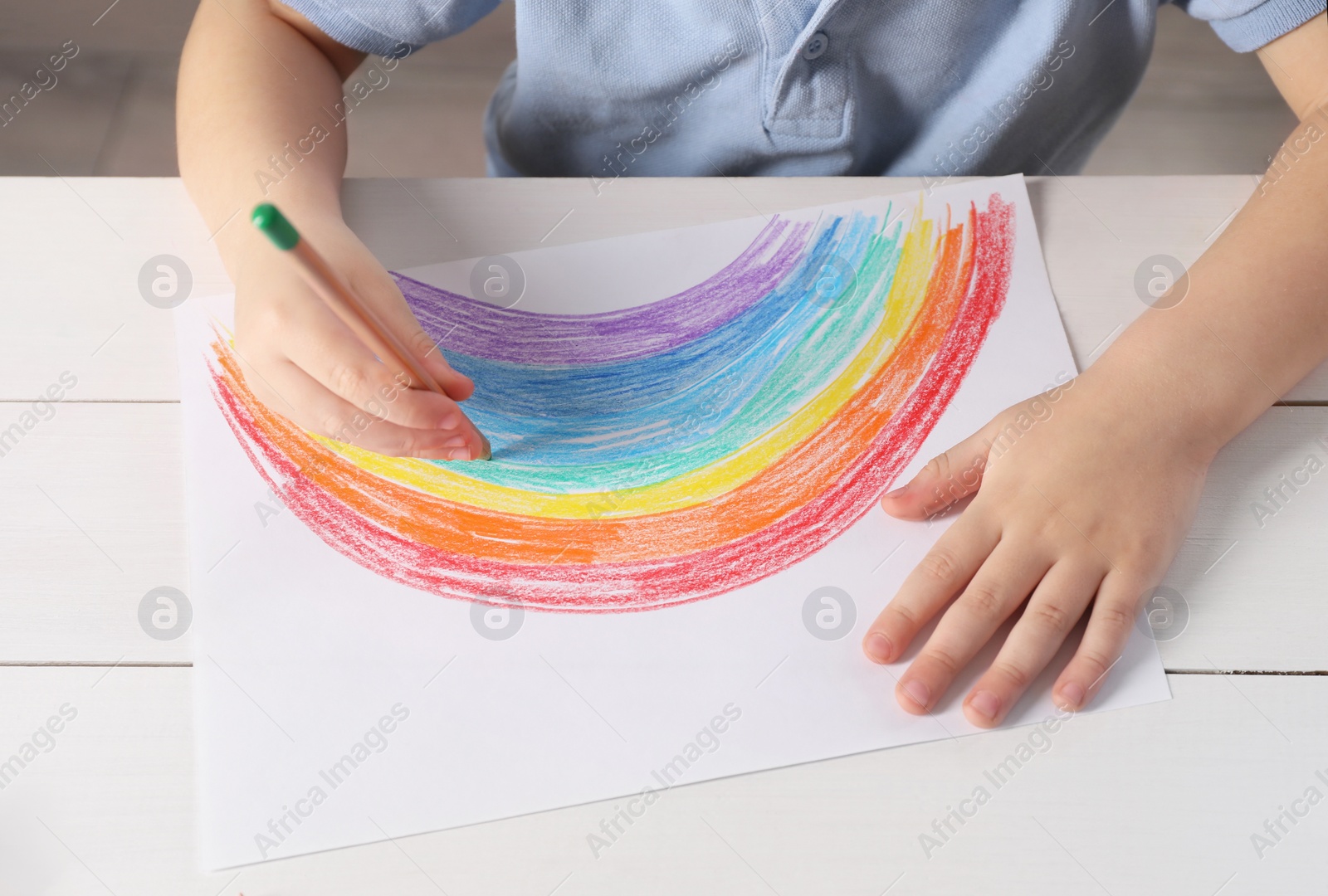 Photo of Little boy drawing rainbow with pencil at white wooden table indoors, closeup. Child`s art