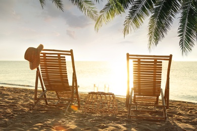 Wooden sun loungers and wicker stand under palm leaves on sandy beach. Summer vacation