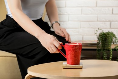 Woman taking mug of coffee from cup coaster at wooden table in room, closeup