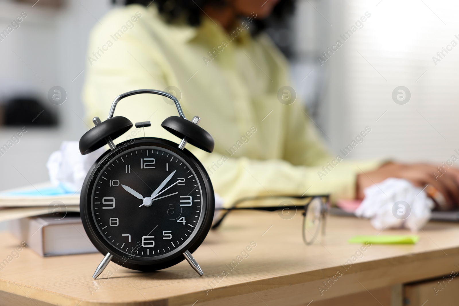 Photo of Deadline concept. Black alarm clock near woman working at wooden desk indoors, selective focus. Space for text