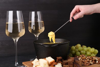 Photo of Woman dipping piece of bread into fondue pot with melted cheese at black wooden table, closeup
