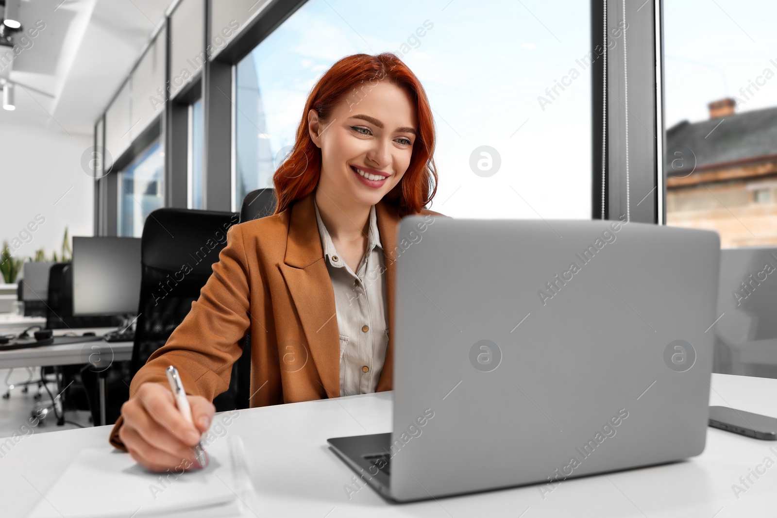 Photo of Happy woman taking notes while working with laptop at white desk in office