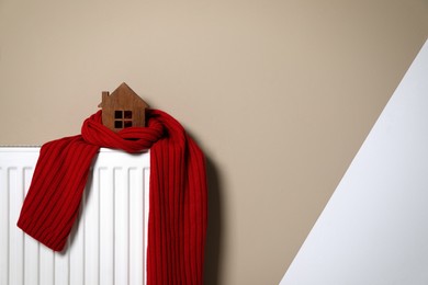 Modern radiator with knitted scarf and wooden house near color wall indoors, space for text. Winter heating efficiency