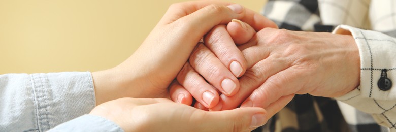 Young and elderly women holding hands on beige background, closeup. Banner design