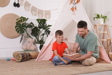 Father and son reading book near toy wigwam at home