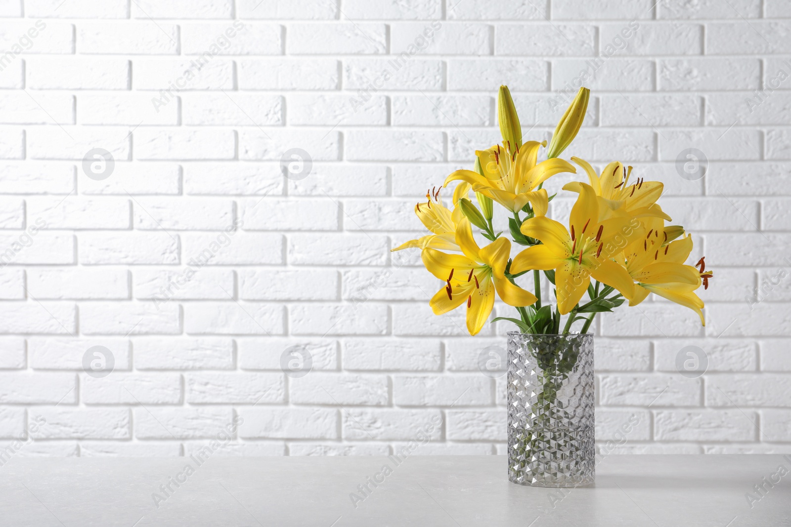 Photo of Glass vase with yellow lily flowers on marble table near white brick wall, space for text