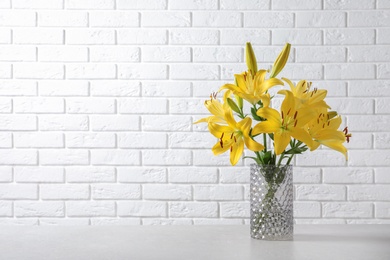 Photo of Glass vase with yellow lily flowers on marble table near white brick wall, space for text