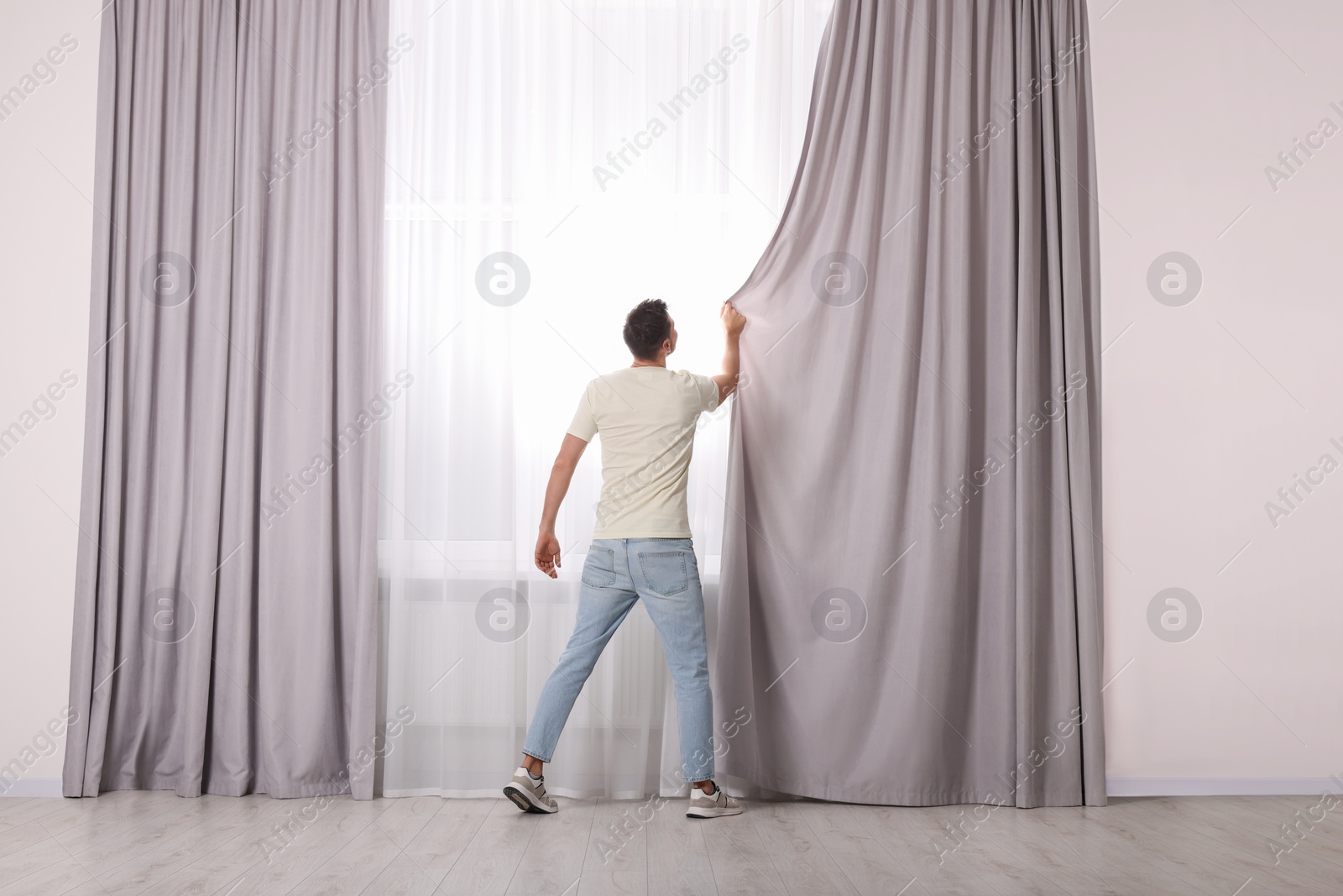 Photo of Man closing window curtains at home, back view