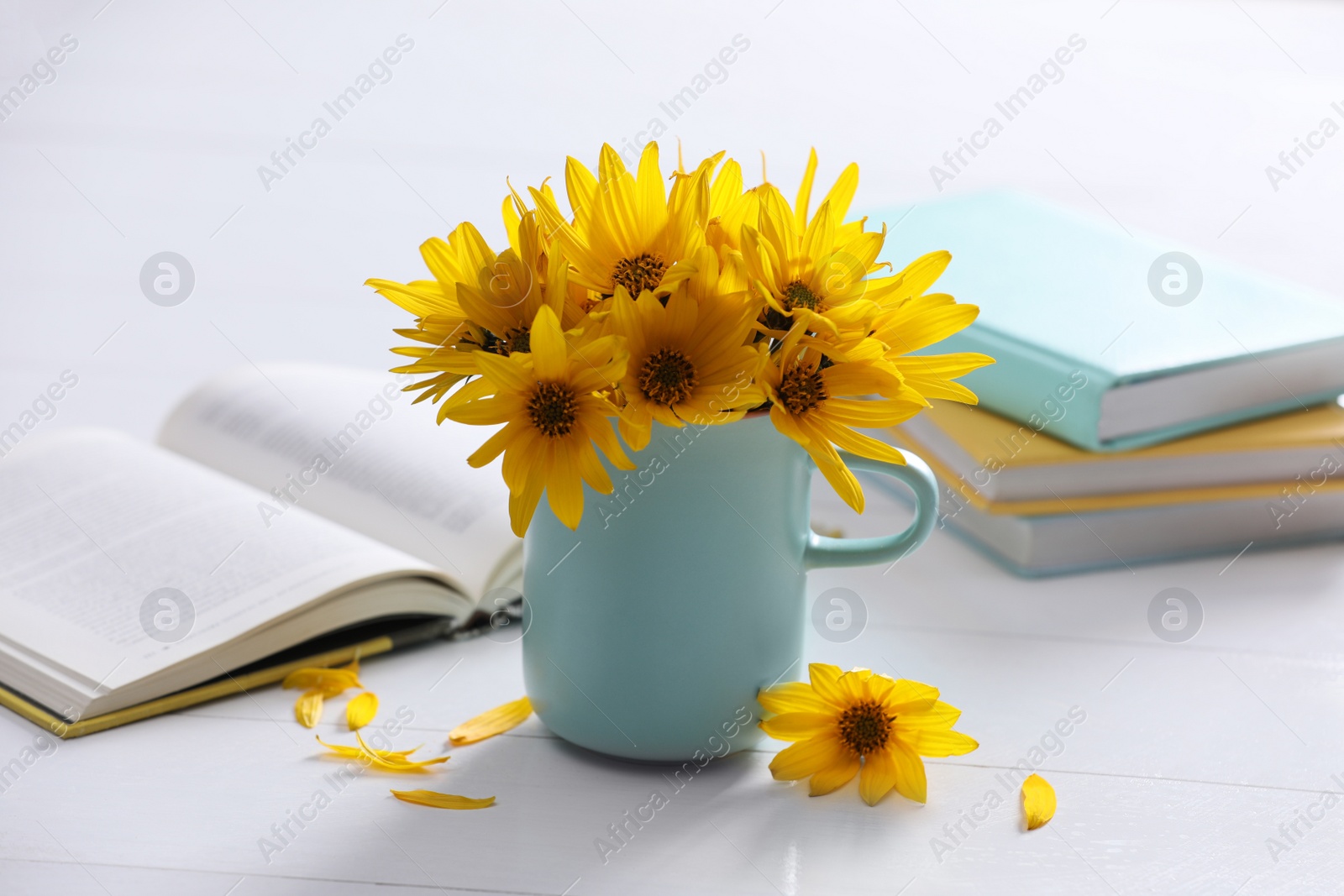 Photo of Beautiful bright yellow flowers in light blue cup near books on white table