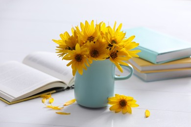 Photo of Beautiful bright yellow flowers in light blue cup near books on white table
