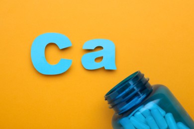 Photo of Open bottle and calcium symbol made of light blue letters on orange background, flat lay