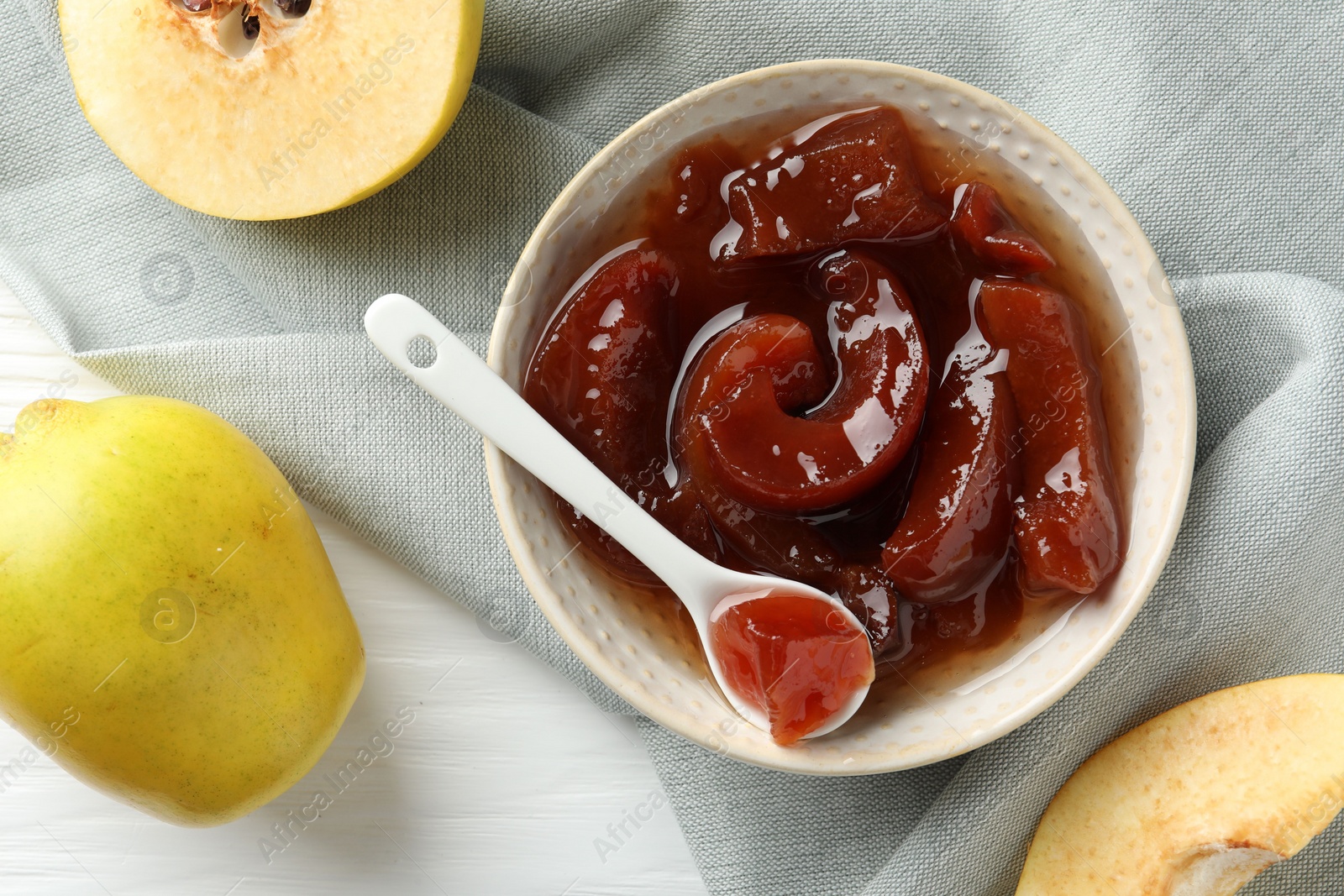 Photo of Tasty homemade quince jam in bowl, spoon and fruits on white wooden table, flat lay