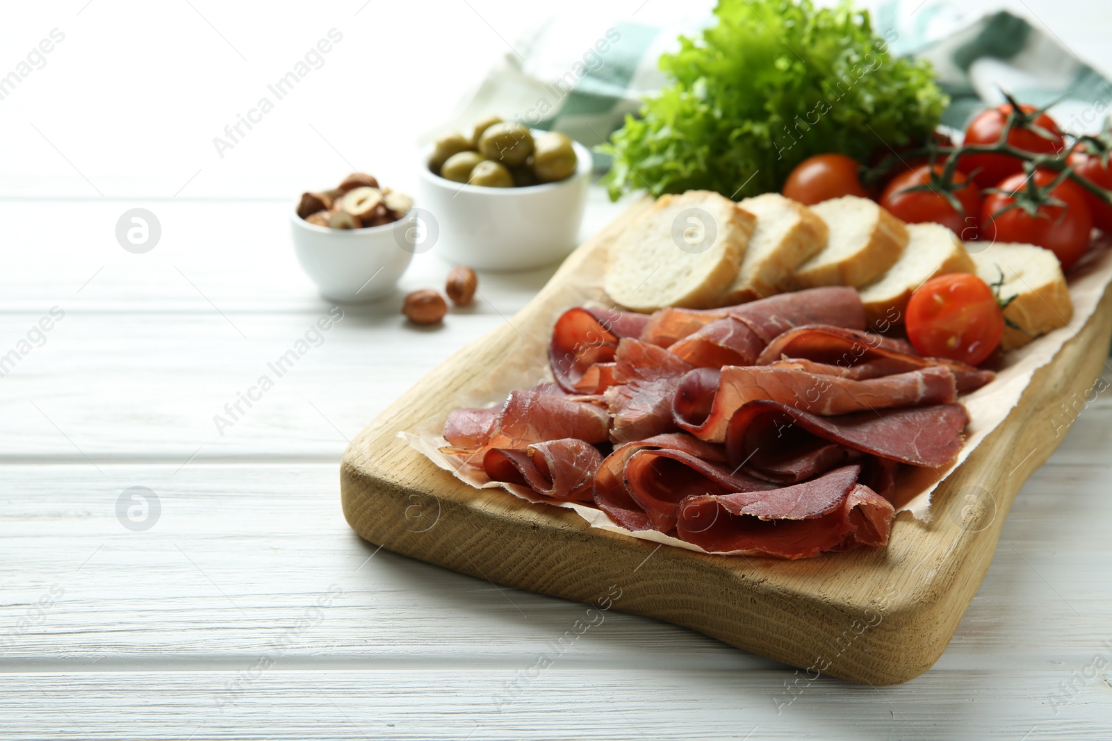 Photo of Board with delicious bresaola served with bread and tomato on white wooden table. Space for text