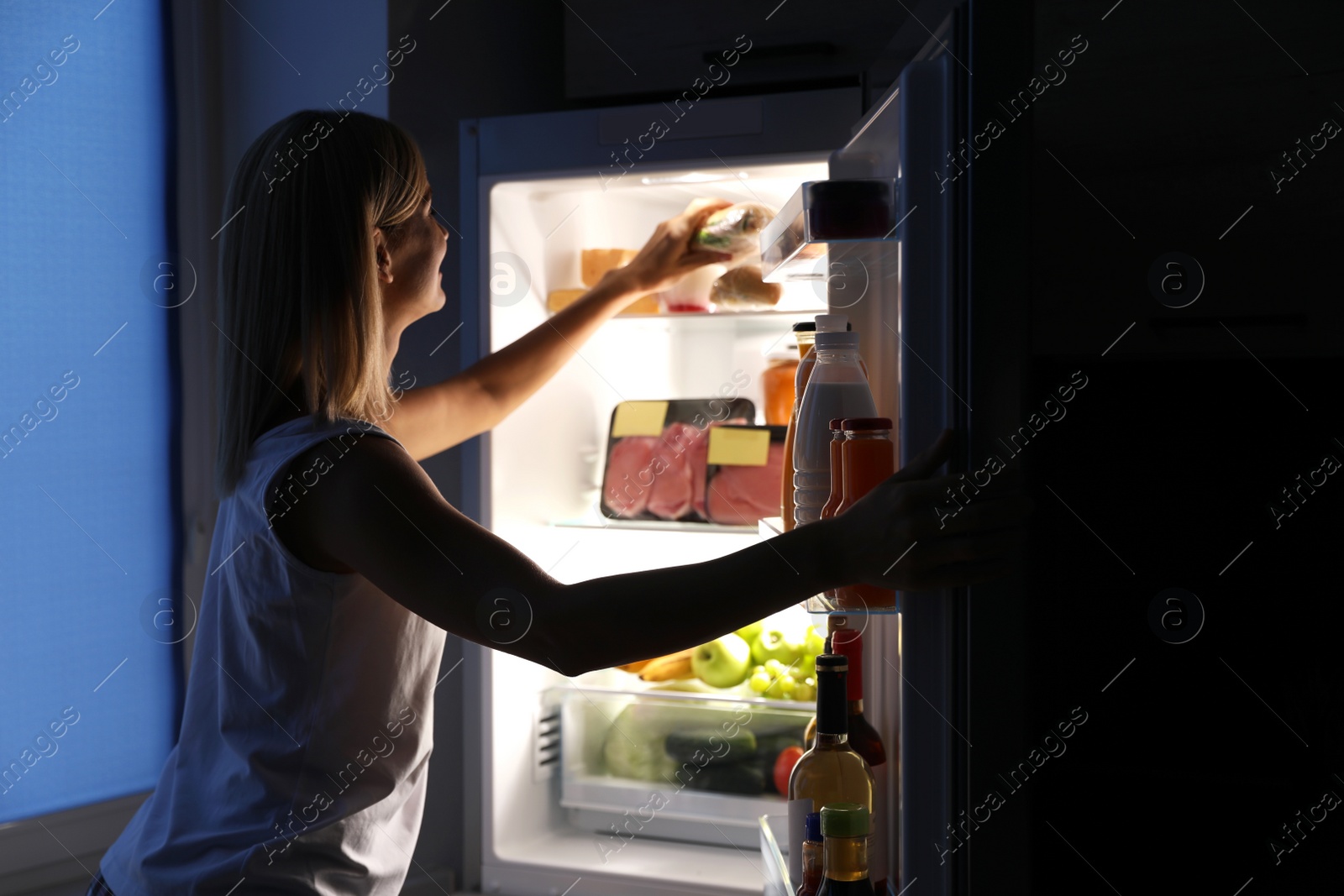 Photo of Woman taking sandwich out of refrigerator in kitchen at night