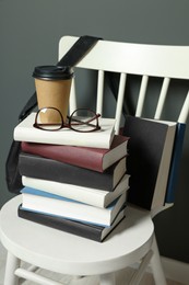 Stack of different books, paper cup and glasses on white chair