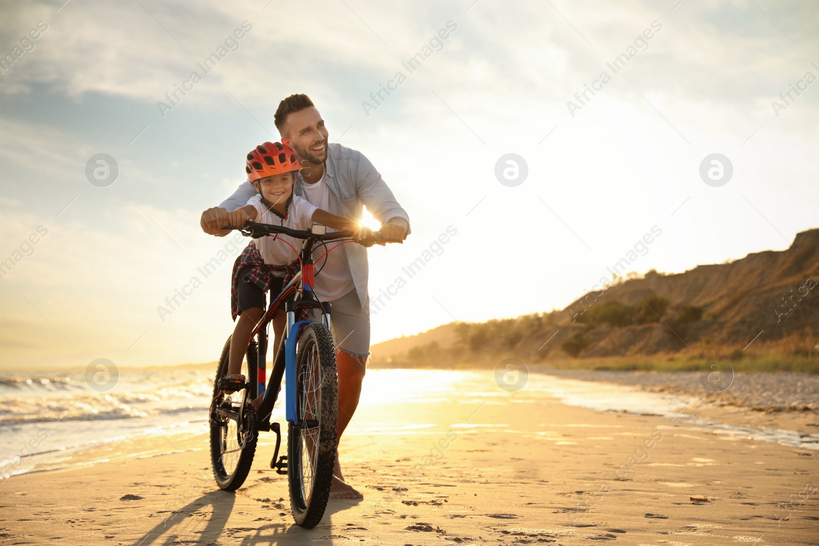 Photo of Happy father teaching son to ride bicycle on sandy beach near sea at sunset