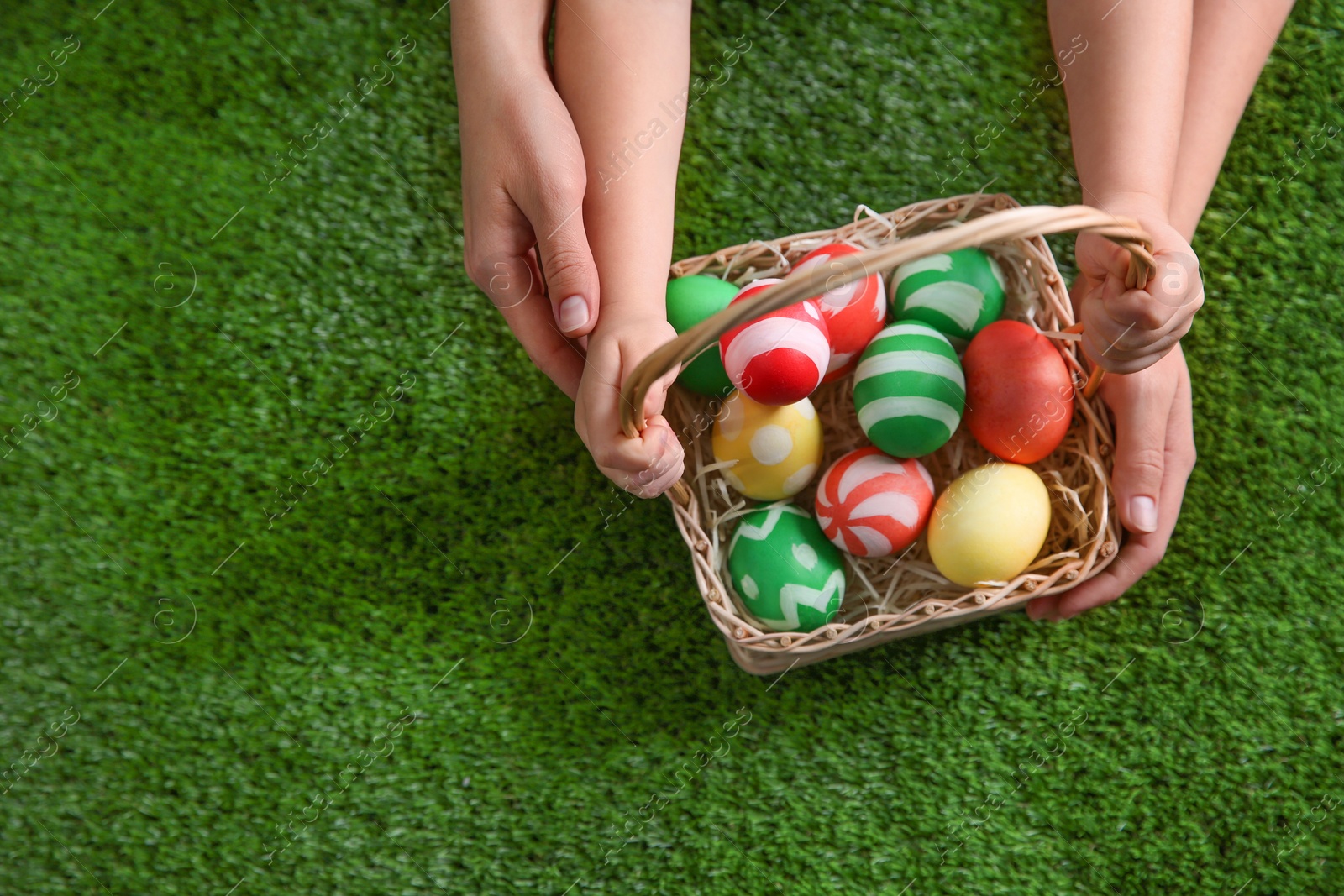 Photo of Mother and her little child with basket of painted Easter eggs on green grass, top view. Space for text