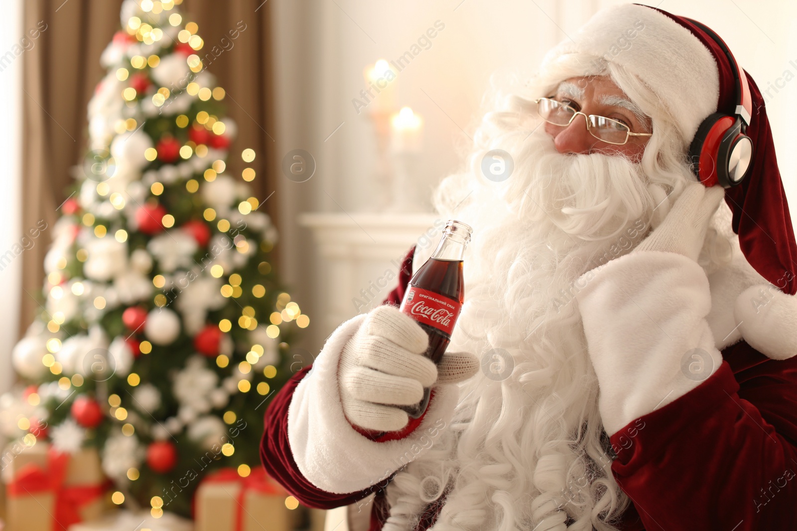 Photo of MYKOLAIV, UKRAINE - JANUARY 18, 2021: Santa Claus holding Coca-Cola bottle and listening to music with headphones in room decorated for Christmas