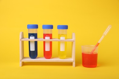 Beaker and test tubes with colorful liquids in wooden stand on yellow background. Kids chemical experiment set