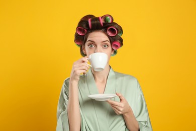 Beautiful young woman in silk bathrobe with hair curlers and cup of drink on yellow background