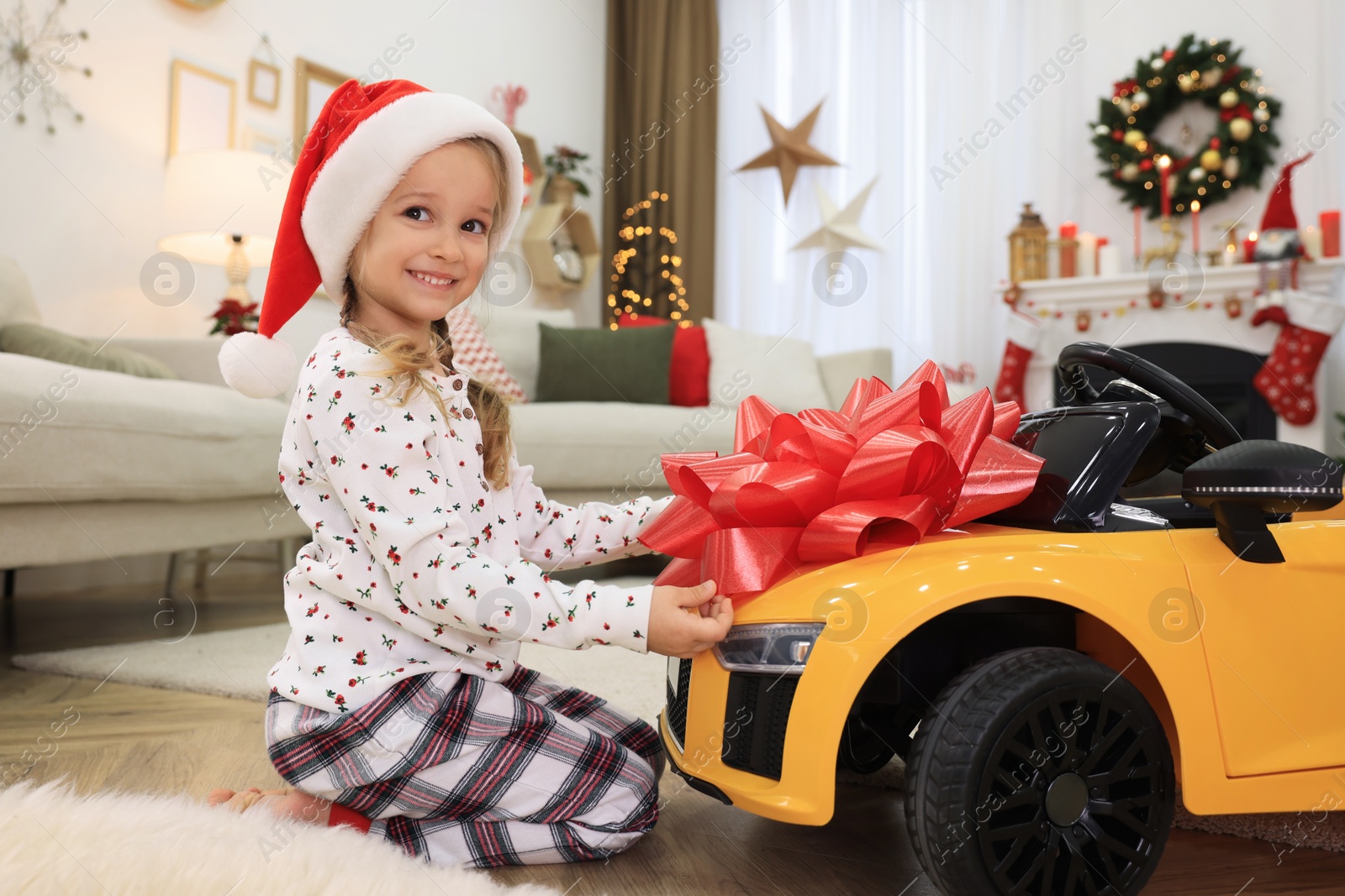 Photo of Cute little girl near toy car with big red bow in room decorated for Christmas