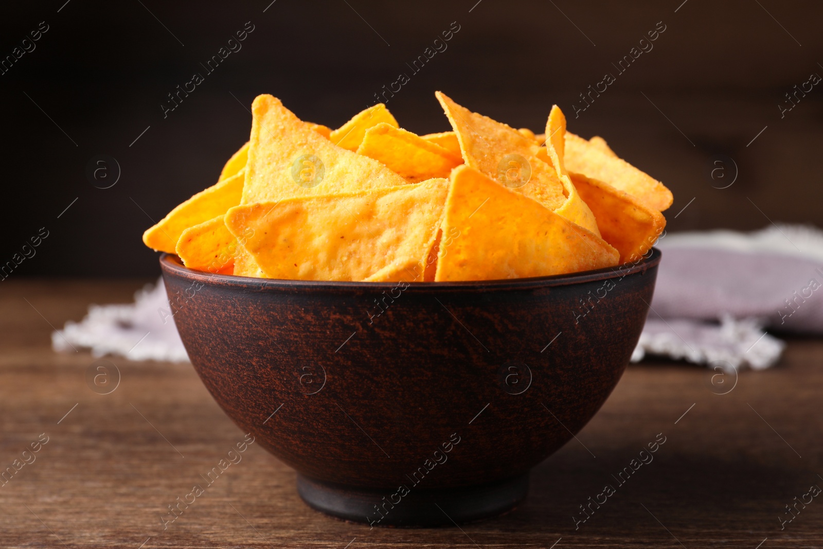 Photo of Tortilla chips (nachos) in bowl on wooden table against dark background, closeup