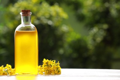Photo of Rapeseed oil in glass bottle and yellow flowers on white wooden table outdoors, space for text