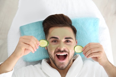 Photo of Young man with clay mask on his face holding cucumber slices in spa salon, above view