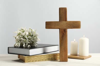 Burning church candles, wooden cross, ecclesiastical books and flowers on white table