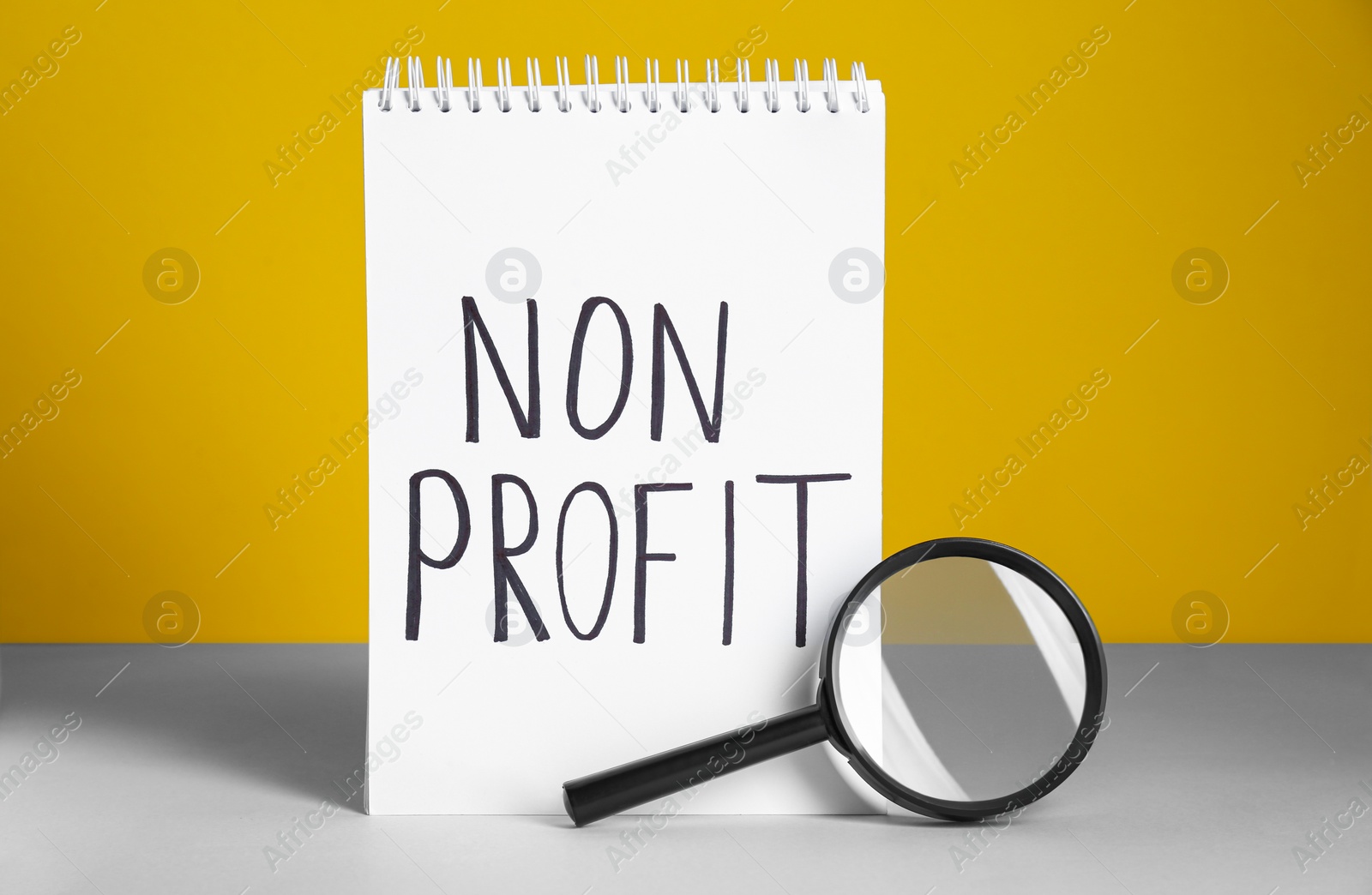 Photo of Notebook with phrase Non Profit and magnifying glass on white table