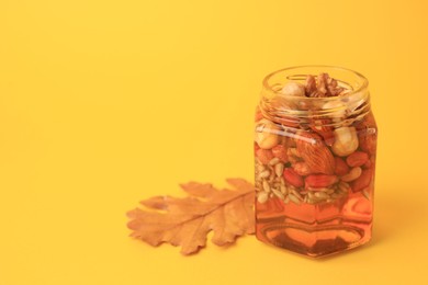 Different nuts with honey in jar and dry leaf on yellow background. Space for text