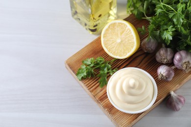 Tasty mayonnaise sauce in bowl, parsley, garlic and lemon on white wooden table, above view. Space for text