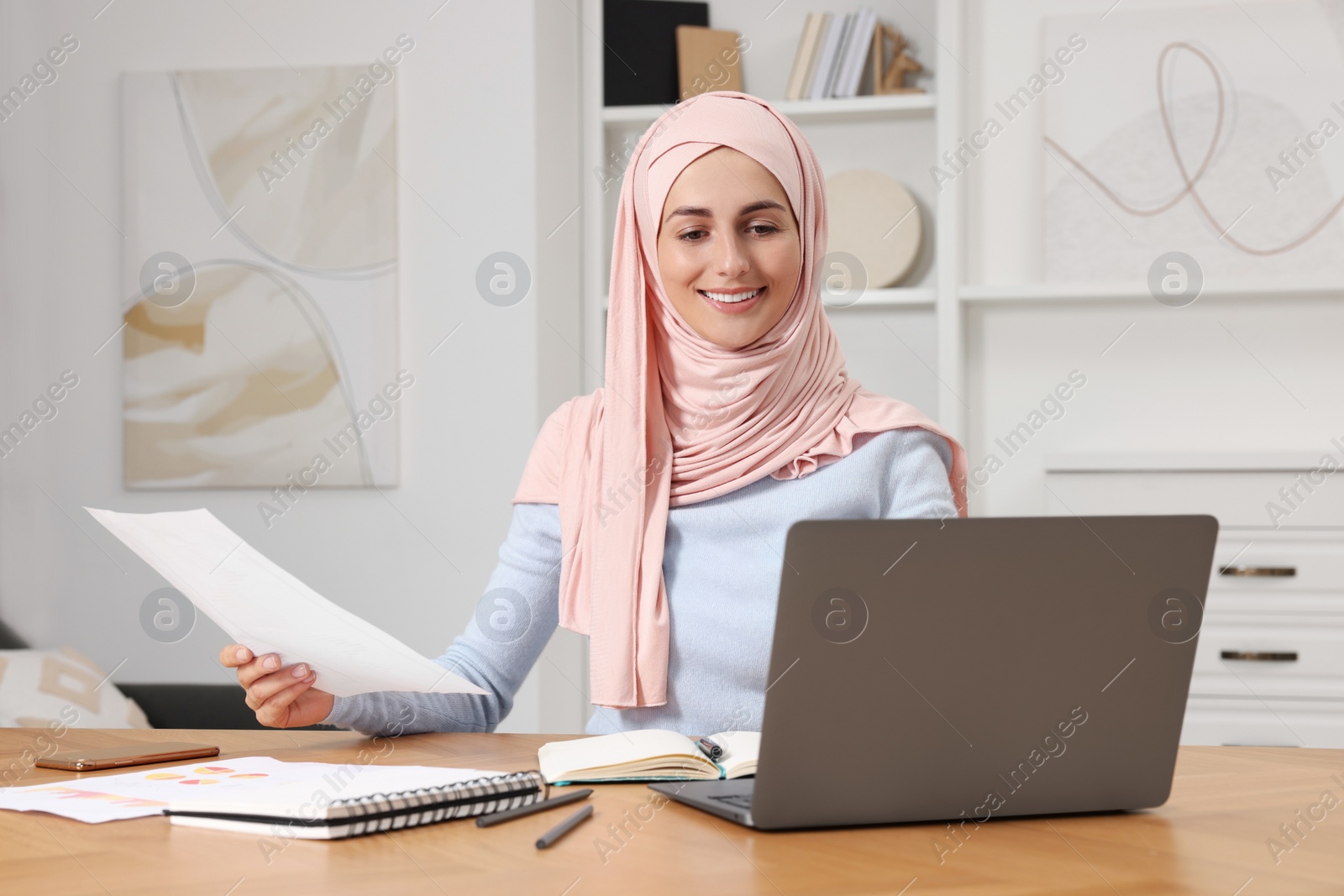 Photo of Muslim woman working near laptop at wooden table in room