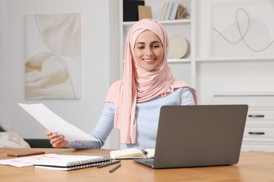 Muslim woman working near laptop at wooden table in room