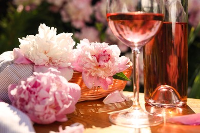 Photo of Bottle and glass of rose wine near beautiful peonies on wooden table in garden, closeup