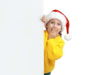 Photo of Emotional child in Santa hat peeping out of blank banner on white background. Christmas celebration