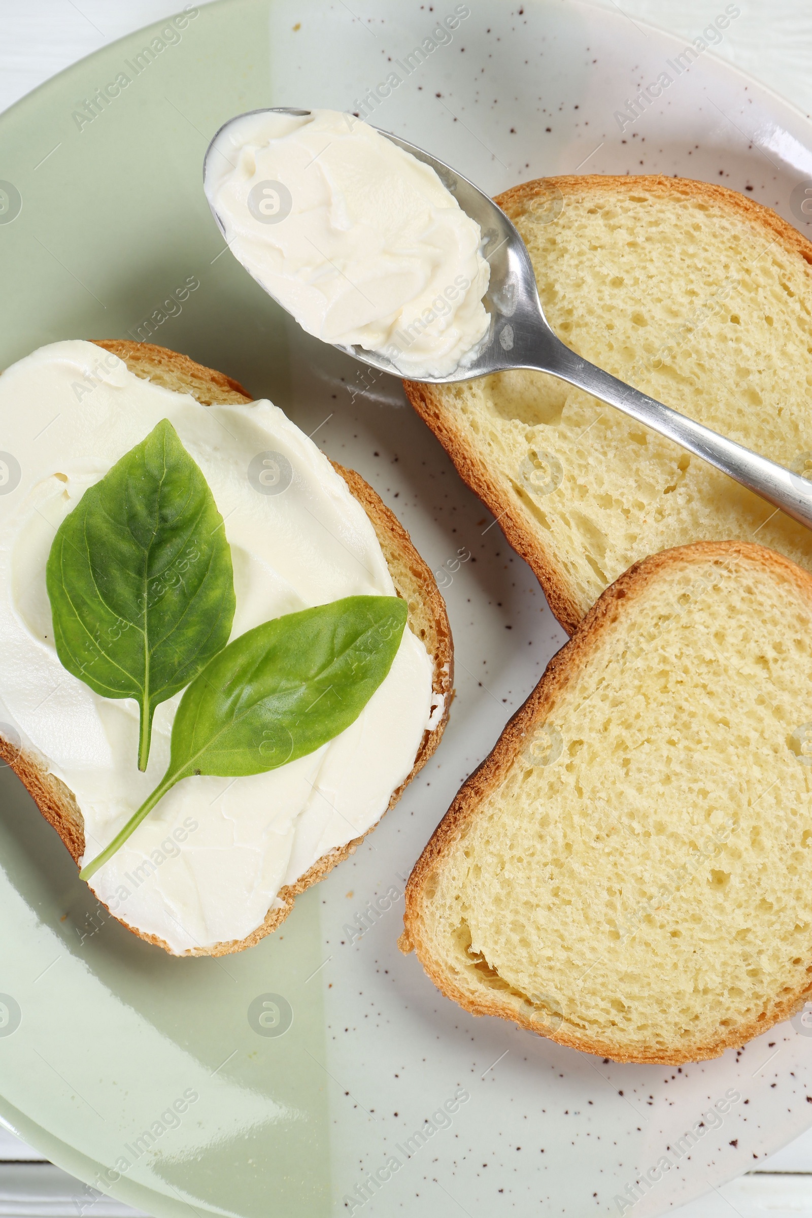 Photo of Pieces of bread with cream cheese and basil leaves on white wooden table, top view