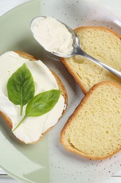 Photo of Pieces of bread with cream cheese and basil leaves on white wooden table, top view