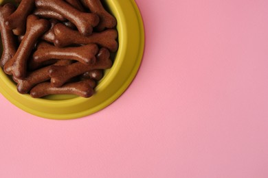 Photo of Yellow bowl with bone shaped dog cookies on pink background, top view. Space for text