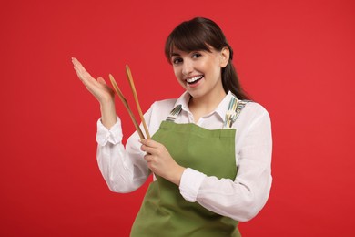 Photo of Happy professional confectioner in apron holding wooden spoon and spatula on red background