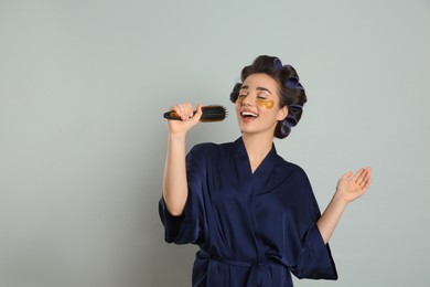 Happy young woman in silk bathrobe with hair curlers and eye patches singing into hairbrush on grey background, space for text