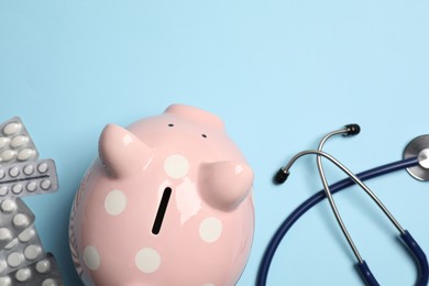 Photo of Piggy bank, stethoscope and pills on light blue background, flat lay. Medical insurance