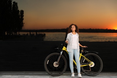 Beautiful young African-American woman with bicycle near glass building at sunset. Space for text