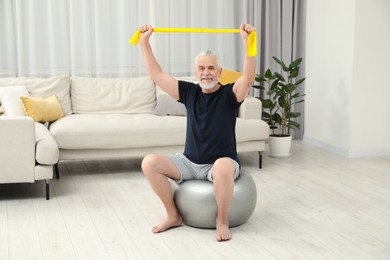 Photo of Senior man doing exercise with elastic resistance band on fitness ball at home